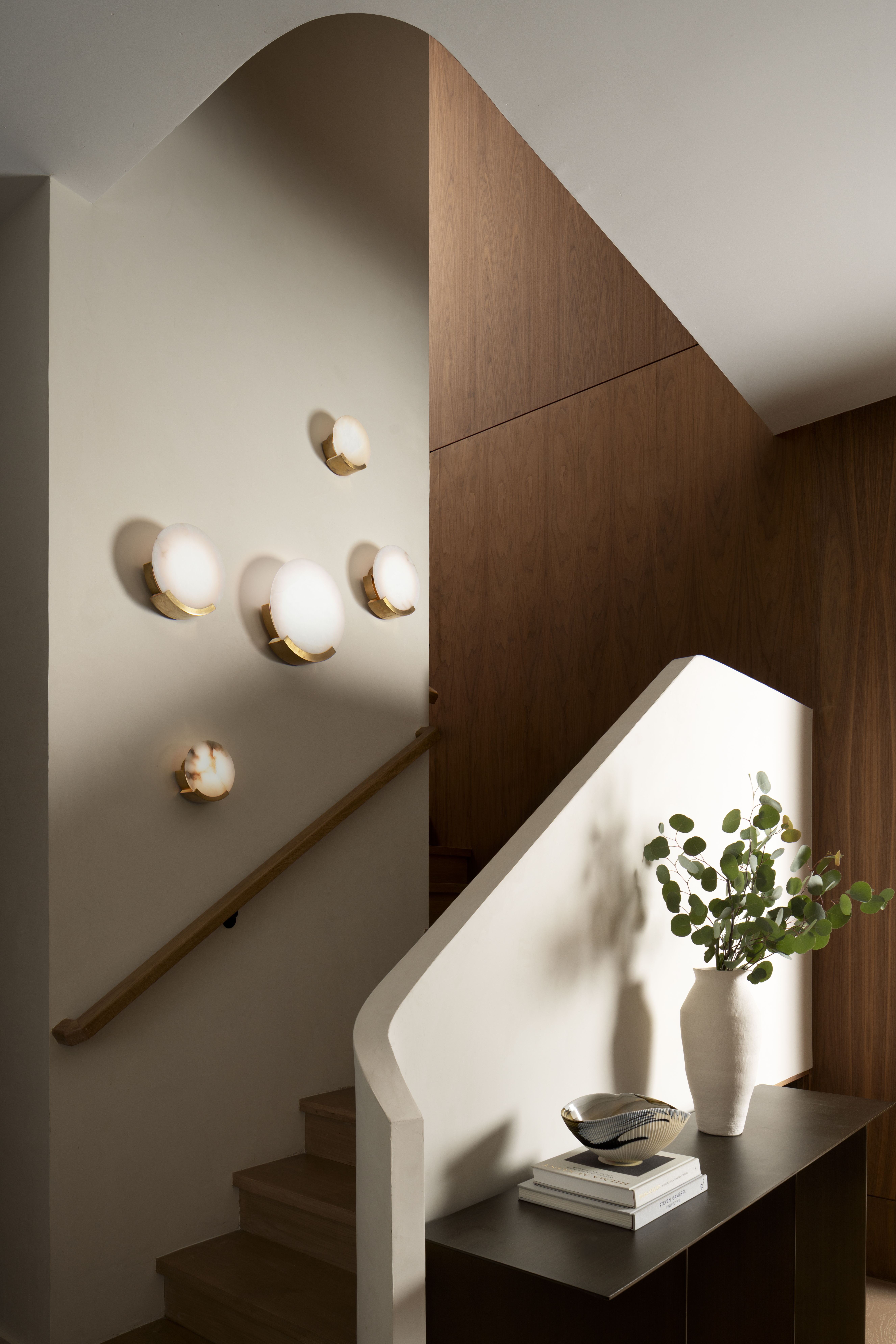 A walnut stair hall with cream walls, globe sconces, and a sunlit white balustrade